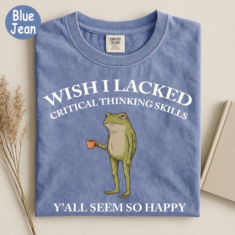 May include: A blue jean colored t-shirt featuring a graphic of a green frog holding a small orange mug. The text above the frog reads WISH I LACKED CRITICAL THINKING SKILLS and the text below the frog reads Y'ALL SEEM SO HAPPY in white font.