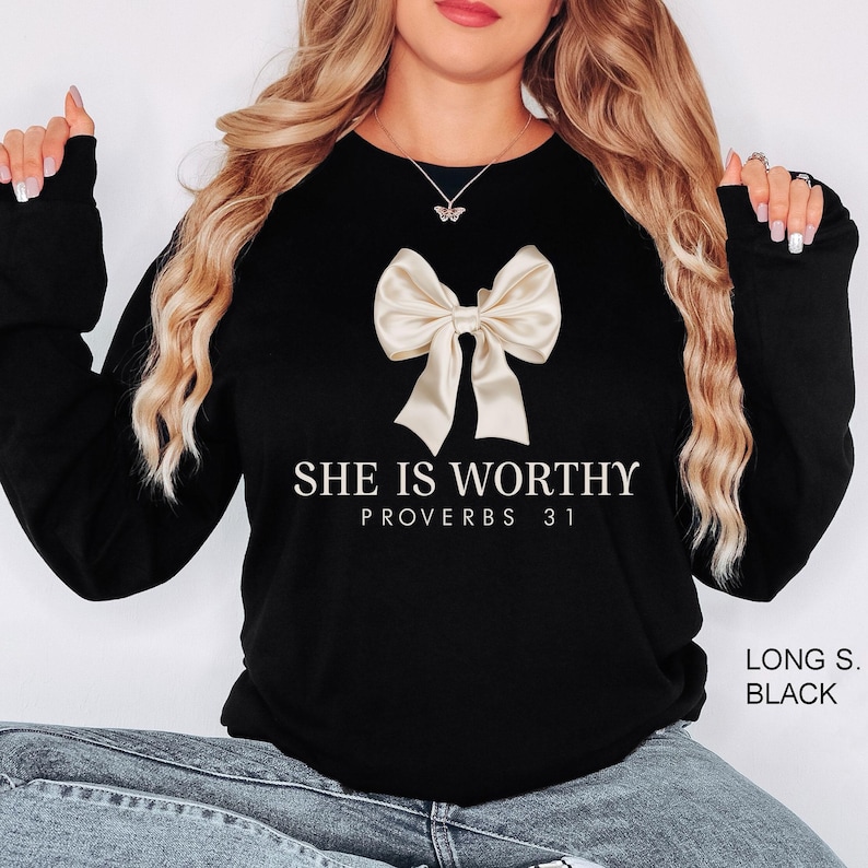 May include: Black long-sleeve sweatshirt with a cream-colored bow graphic. The text "SHE IS WORTHY PROVERBS 31" is printed below the bow. The sweatshirt is a solid black color.
