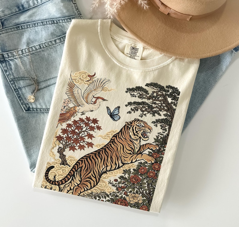 May include: A cream-colored t-shirt featuring a graphic print of a tiger, a flying crane, a blue butterfly, and floral elements. The shirt has a crew neckline and a Comfort Colors brand label on the inside collar. The design includes stylized trees.