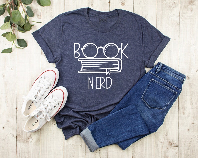 May include: Navy blue t-shirt with the words "BOOK NERD" in white, featuring a book and glasses design. The shirt is paired with blue jeans and white sneakers with red accents. The items are arranged on a light wood-grain surface.