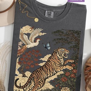 May include: A charcoal gray crew neck t-shirt featuring a graphic print of a leaping tiger, a flying crane, a blue butterfly, stylized clouds, and floral elements. The shirt has a Comfort Colors label at the neck and the word Pepper printed below.
