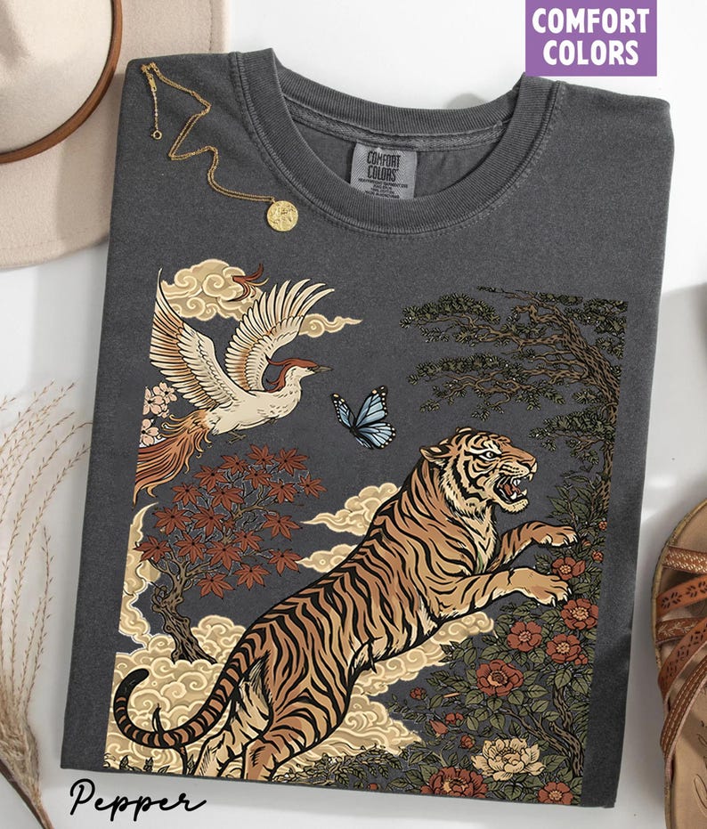 May include: A charcoal gray crew neck t-shirt featuring a graphic print of a leaping tiger, a flying crane, a blue butterfly, stylized clouds, and floral elements. The shirt has a Comfort Colors label at the neck and the word Pepper printed below.