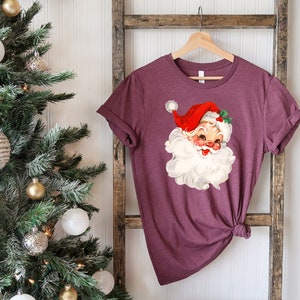 May include: Maroon t-shirt featuring a vintage Santa Claus graphic. The Santa illustration has a red hat, white beard, and rosy cheeks. The shirt is displayed on a wooden ladder with a Christmas tree in the background, decorated with ornaments.