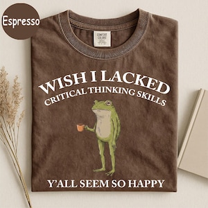 May include: A brown crew neck t-shirt featuring a graphic of a green frog holding a small brown cup. The text reads WISH I LACKED CRITICAL THINKING SKILLS Y'ALL SEEM SO HAPPY in white capital letters. The shirt has a Comfort Colors brand label.