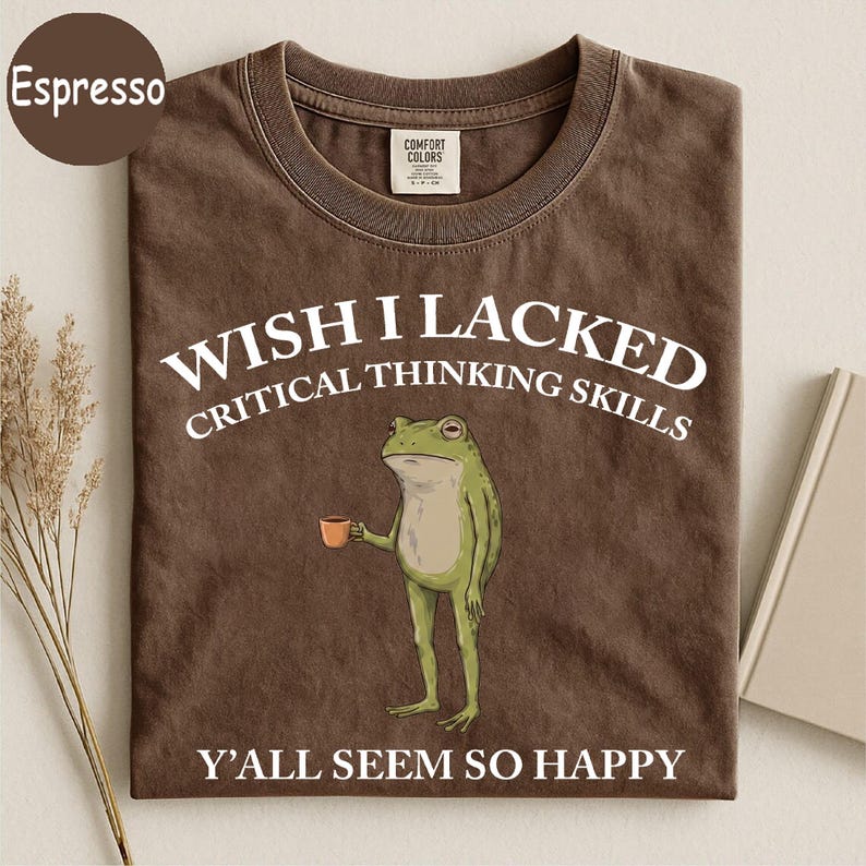 May include: A brown crew neck t-shirt featuring a graphic of a green frog holding a small brown cup. The text reads WISH I LACKED CRITICAL THINKING SKILLS Y'ALL SEEM SO HAPPY in white capital letters. The shirt has a Comfort Colors brand label.