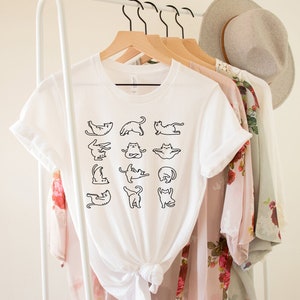 Op de afbeelding: Wit T-shirt met een zwarte omtrekgrafiek van katten die yoga-houdingen aannemen. Het shirt hangt aan een hanger met andere kleding.