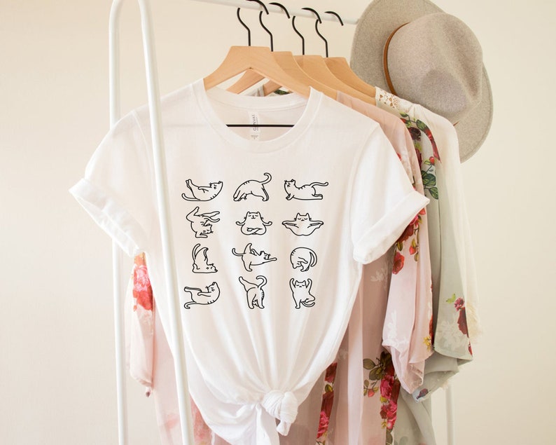 Op de afbeelding: Wit T-shirt met een zwarte omtrekgrafiek van katten die yoga-houdingen aannemen. Het shirt hangt aan een hanger met andere kleding.