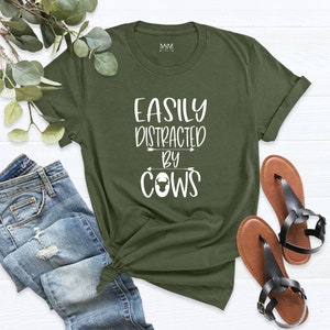 May include: Olive green t-shirt with the text "EASILY DISTRACTED BY COWS" in white. The shirt is paired with blue jeans and brown sandals with black straps. A sprig of greenery is in the background.