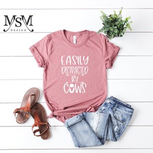 May include: A dusty rose t-shirt with the text "EASILY DISTRACTED BY COWS" in white. The shirt is paired with blue jeans and brown sandals. A small potted plant is also in the image.