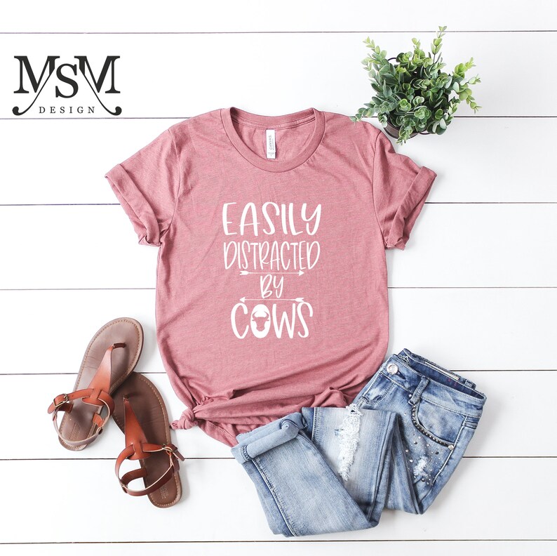 May include: A dusty rose t-shirt with the text "EASILY DISTRACTED BY COWS" in white. The shirt is paired with blue jeans and brown sandals. A small potted plant is also in the image.