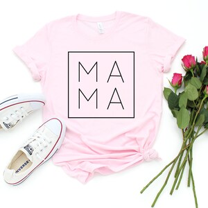 May include: A light pink t-shirt with the word "MAMA" in a black square. White sneakers and a bouquet of pink roses are arranged on a white surface. The shirt is folded at the bottom.