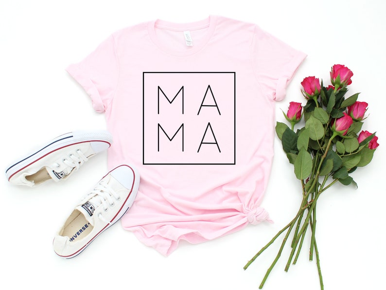 May include: A light pink t-shirt with the word "MAMA" in a black square. White sneakers and a bouquet of pink roses are arranged on a white surface. The shirt is folded at the bottom.