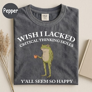 May include: A charcoal gray crew neck t-shirt featuring a graphic of a green frog holding a small orange mug. The text reads: WISH I LACKED CRITICAL THINKING SKILLS Y'ALL SEEM SO HAPPY. The shirt has a Comfort Colors brand label at the inner neckline.