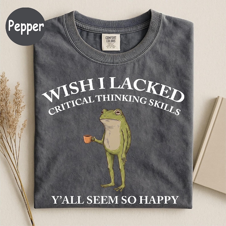May include: A charcoal gray crew neck t-shirt featuring a graphic of a green frog holding a small orange mug. The text reads: WISH I LACKED CRITICAL THINKING SKILLS Y'ALL SEEM SO HAPPY. The shirt has a Comfort Colors brand label at the inner neckline.