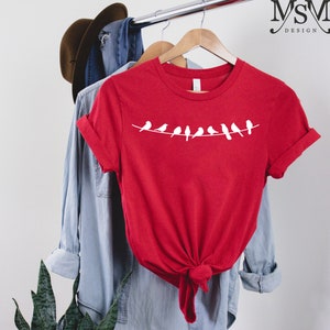 May include: Red t-shirt with a white graphic of a line of birds on a wire. The shirt is hanging on a wooden hanger.