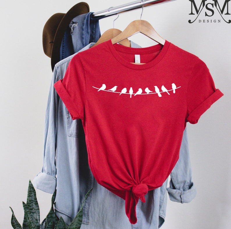 May include: Red t-shirt with a white graphic of a line of birds on a wire. The shirt is hanging on a wooden hanger.