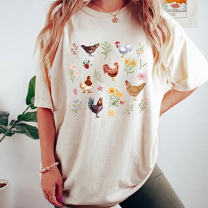 May include: A cream-colored t-shirt with a graphic print of chickens and flowers. The graphic features a variety of chickens in different colors and poses, surrounded by colorful flowers and leaves.