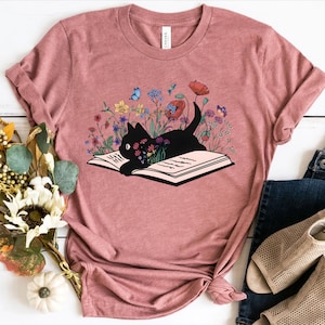 Pu&ograve; includere: Una maglietta rosa con un gatto nero che legge un libro circondato da fiori colorati. Il gatto &egrave; sdraiato sul libro aperto con la testa appoggiata sulle pagine. I fiori sono di vari colori, tra cui rosso, rosa, giallo e blu.