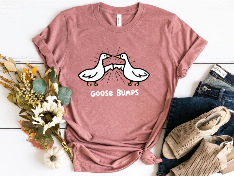 Puede incluir: Una camiseta rosa con un gr&aacute;fico de dos gansos que se miran con el texto "Goose Bumps" debajo.
