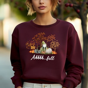 May include: A maroon long-sleeve sweatshirt featuring a graphic print of a gnome sitting on pumpkins with a black cat, autumn trees, and the white text "Ahhhh...fall" printed below the illustration on the front of the garment.