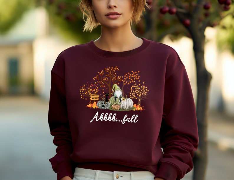May include: A maroon long-sleeve sweatshirt featuring a graphic print of a gnome sitting on pumpkins with a black cat, autumn trees, and the white text "Ahhhh...fall" printed below the illustration on the front of the garment.