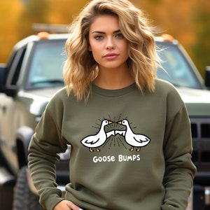 Puede incluir: Una sudadera verde con un gr&aacute;fico blanco de dos gansos que se miran con el texto "Goose Bumps" debajo.