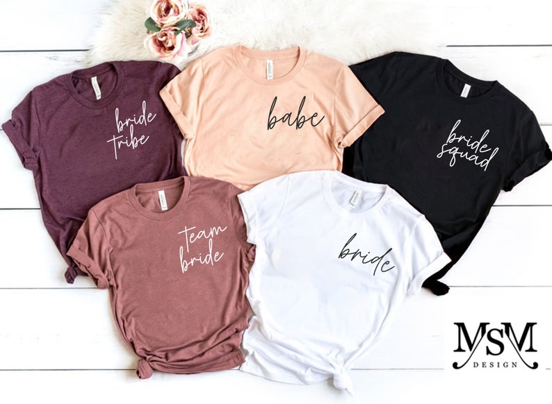 K&ouml;nnte beinhalten: F&uuml;nf kurz&auml;rmelige T-Shirts in verschiedenen Farben: Burgunderrot, Pfirsich, Schwarz, Mauve und Wei&szlig;. Jedes Shirt hat einen anderen Schriftzug: "bride tribe", "babe", "bride squad", "team bride" und "bride". Die Shirts liegen auf einer wei&szlig;en Oberfl&auml;che.