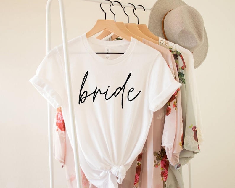May include: White t-shirt with the word "bride" in black script. The shirt is knotted at the waist and hangs on a wooden hanger. Other clothing items and a hat are also on the rack.