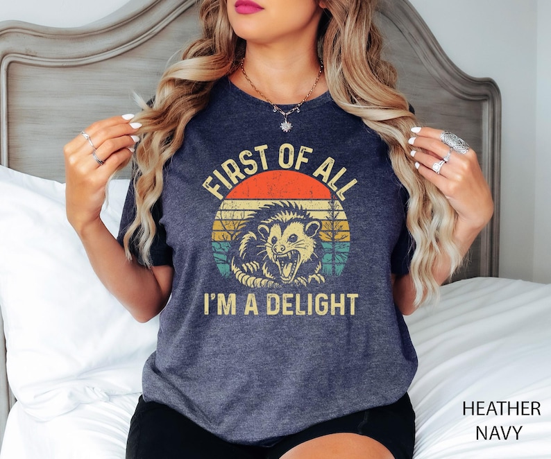 May include: A dark heather navy blue t-shirt with a vintage-style graphic of a possum with the text "First of all I'm a delight".