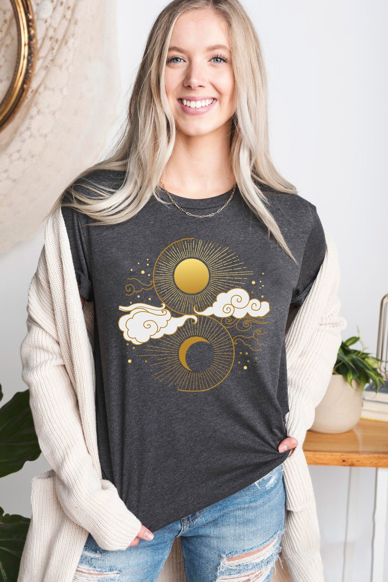 May include: A dark grey t-shirt with a gold sun and moon design. The design features a sun with rays, a crescent moon, and clouds.