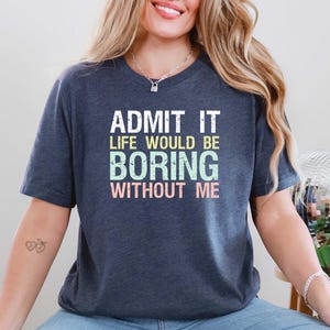 May include: Dark blue t-shirt with the text "ADMIT IT LIFE WOULD BE BORING WITHOUT ME" in white, green, and pink. The shirt has a crew neck and short sleeves. The text is centered on the front of the shirt.