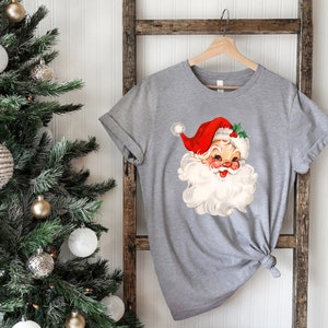 May include: A gray t-shirt featuring a vintage illustration of Santa Claus with a red hat, white beard, and rosy cheeks. The Santa Claus graphic is centered on the shirt, which is hanging on a wooden hanger. The shirt is tied in a knot at the bottom.