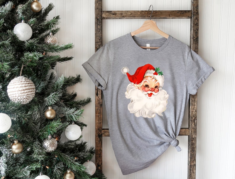May include: A gray t-shirt featuring a vintage illustration of Santa Claus with a red hat, white beard, and rosy cheeks. The Santa Claus graphic is centered on the shirt, which is hanging on a wooden hanger. The shirt is tied in a knot at the bottom.