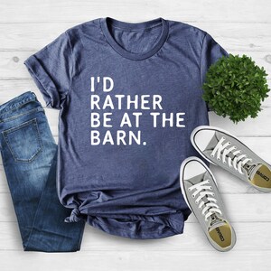 Puede incluir: Una camiseta azul marino con el texto "I'D RATHER BE AT THE BARN." se muestra con vaqueros azules y zapatillas grises. La camiseta est&aacute; hecha de material suave y es adecuada para ropa casual.