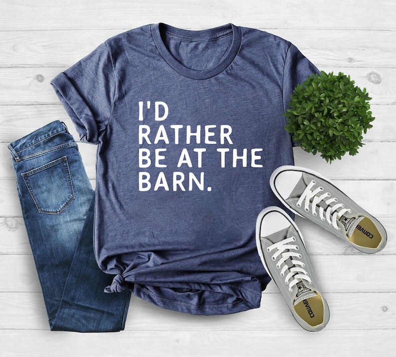 Puede incluir: Una camiseta azul marino con el texto "I'D RATHER BE AT THE BARN." se muestra con vaqueros azules y zapatillas grises. La camiseta est&aacute; hecha de material suave y es adecuada para ropa casual.