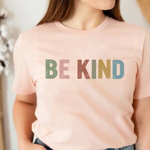 May include: A light pink t-shirt with the words "BE KIND" printed across the front in colorful, textured letters. The letters are in shades of green, pink, red, yellow, and blue. The shirt has short sleeves and a crew neck.