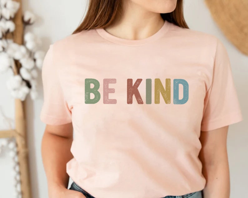 May include: A light pink t-shirt with the words "BE KIND" printed across the front in colorful, textured letters. The letters are in shades of green, pink, red, yellow, and blue. The shirt has short sleeves and a crew neck.