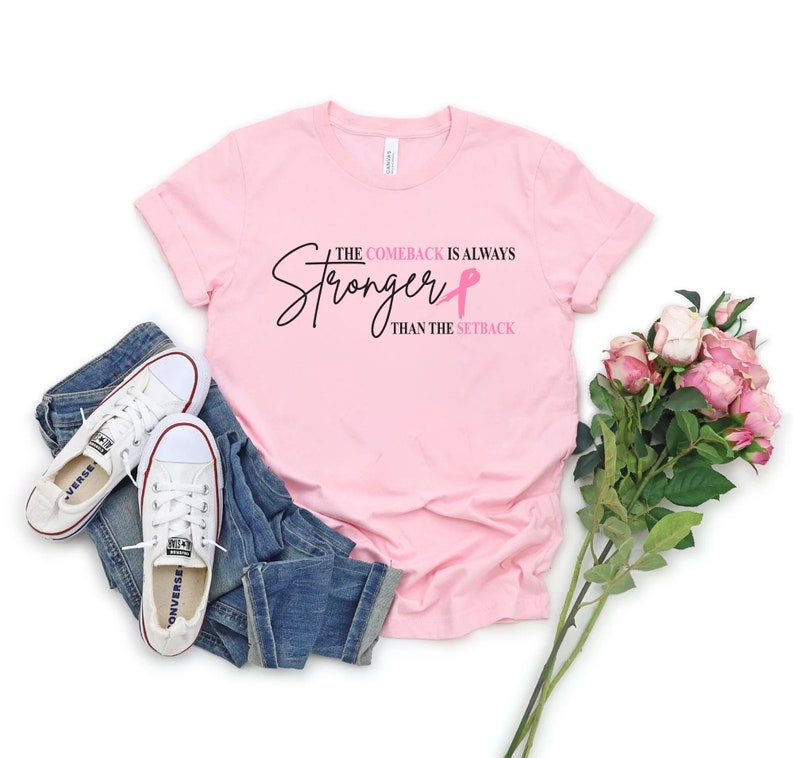 May include: A light pink t-shirt with the text "The Comeback is Always Stronger Than The Setback" in black script and a pink ribbon. The shirt is paired with blue jeans and white Converse sneakers.