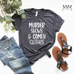 May include: Dark gray t-shirt with the text "MURDER SHOWS & COMFY CLOTHES" in white. The shirt is styled with blue jeans and brown sandals. The shirt is knotted at the waist.