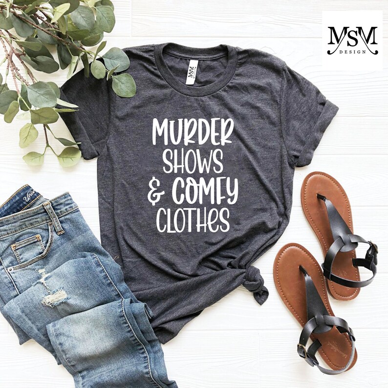 May include: Dark gray t-shirt with the text "MURDER SHOWS & COMFY CLOTHES" in white. The shirt is styled with blue jeans and brown sandals. The shirt is knotted at the waist.
