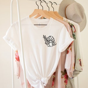 Peut inclure: T-shirt blanc avec un motif de champignon noir. Le motif repr&eacute;sente deux champignons et des feuilles. Le t-shirt est suspendu &agrave; un cintre en bois avec d'autres v&ecirc;tements et un chapeau en arri&egrave;re-plan.
