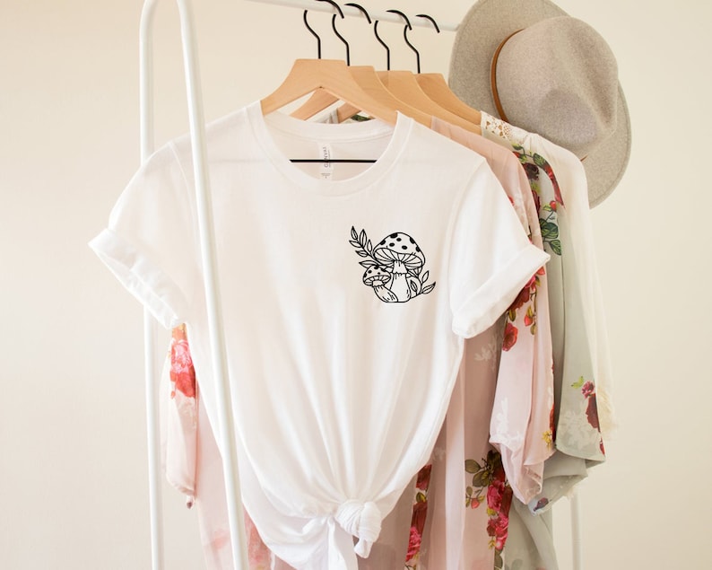 Peut inclure: T-shirt blanc avec un motif de champignon noir. Le motif repr&eacute;sente deux champignons et des feuilles. Le t-shirt est suspendu &agrave; un cintre en bois avec d'autres v&ecirc;tements et un chapeau en arri&egrave;re-plan.