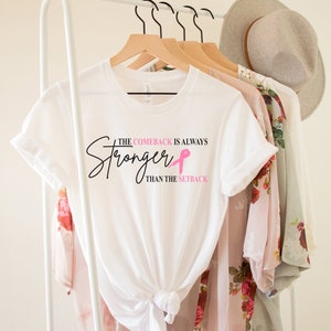 May include: White t-shirt with the text "The Comeback is Always Stronger Than The Setback" in black script and a pink ribbon. The shirt is hanging on a wooden hanger with other clothing items and a hat.