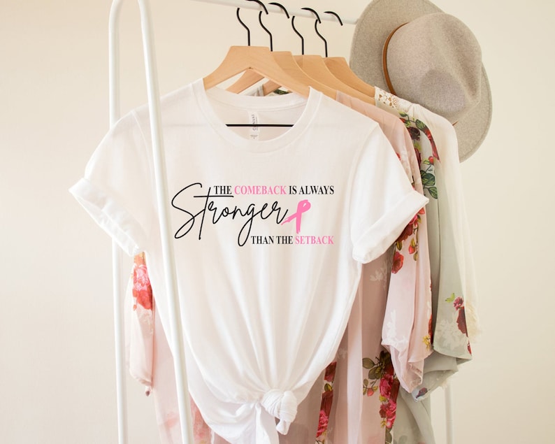 May include: White t-shirt with the text "The Comeback is Always Stronger Than The Setback" in black script and a pink ribbon. The shirt is hanging on a wooden hanger with other clothing items and a hat.