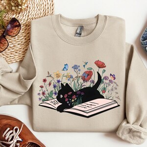 May include: A beige sweatshirt with a black cat reading a book surrounded by colorful flowers and butterflies. The cat is lying on an open book with pink pages.