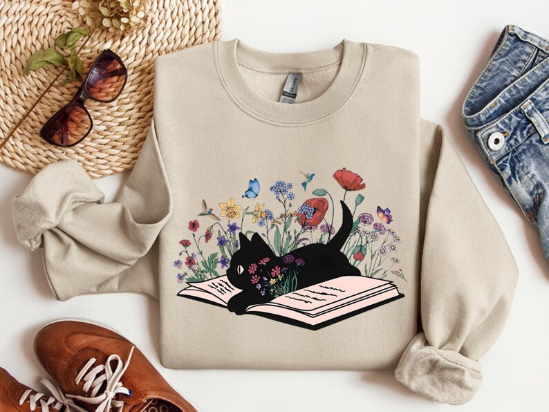 May include: A beige sweatshirt with a black cat reading a book surrounded by colorful flowers and butterflies. The cat is lying on an open book with pink pages.
