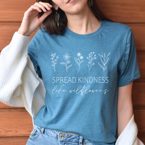 Op de afbeelding: Een tealblauw t-shirt met een witte afbeelding van wilde bloemen en de tekst "SPREAD KINDNESS like wildflowers".