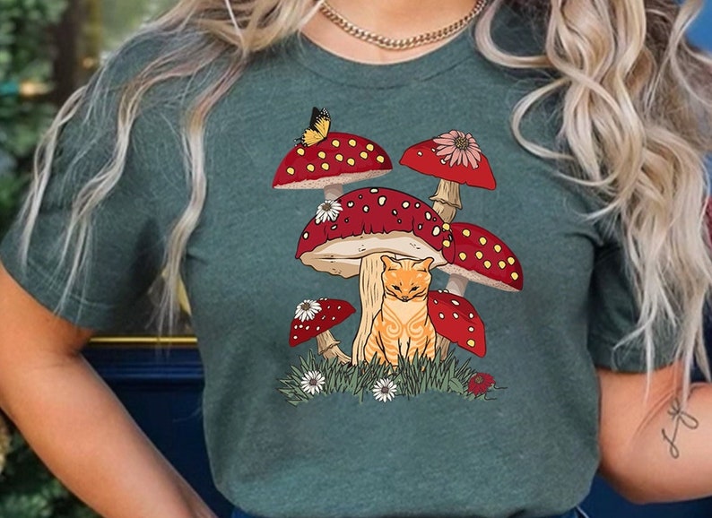 K&ouml;nnte beinhalten: Dunkelgr&uuml;nes T-Shirt mit einer skurrilen Illustration einer rot getigerten Katze, die zwischen roten Pilzen mit wei&szlig;en Punkten sitzt. Ein Schmetterling sitzt auf einem Pilz, umgeben von G&auml;nsebl&uuml;mchen und Gras.