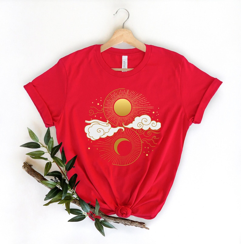 May include: Red t-shirt featuring a gold sun and moon design. The sun and moon are surrounded by clouds and stars.