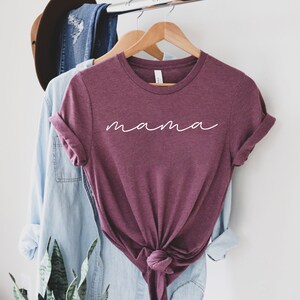 Puede incluir: Una camiseta granate con la palabra "mama" en escritura blanca. La camiseta est&aacute; colgada en una percha de madera, con una camisa abotonada azul claro y vaqueros en el fondo. Las mangas est&aacute;n enrolladas.